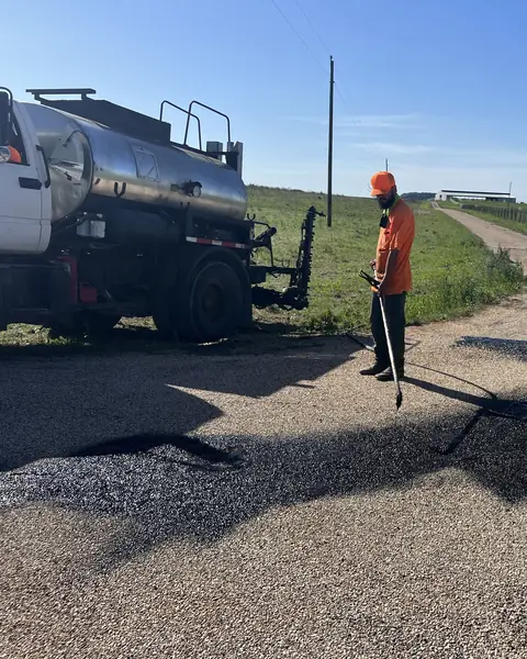 Crew member spraying hot asphalt binder from a distributor truck on a rural drive